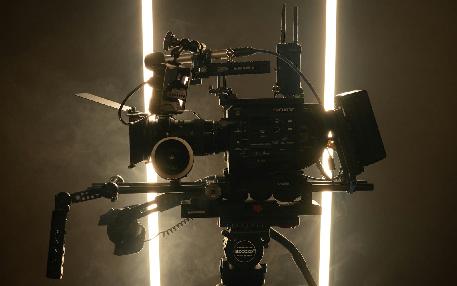 A rigged cinema camera silhouetted against two light tubes in haze