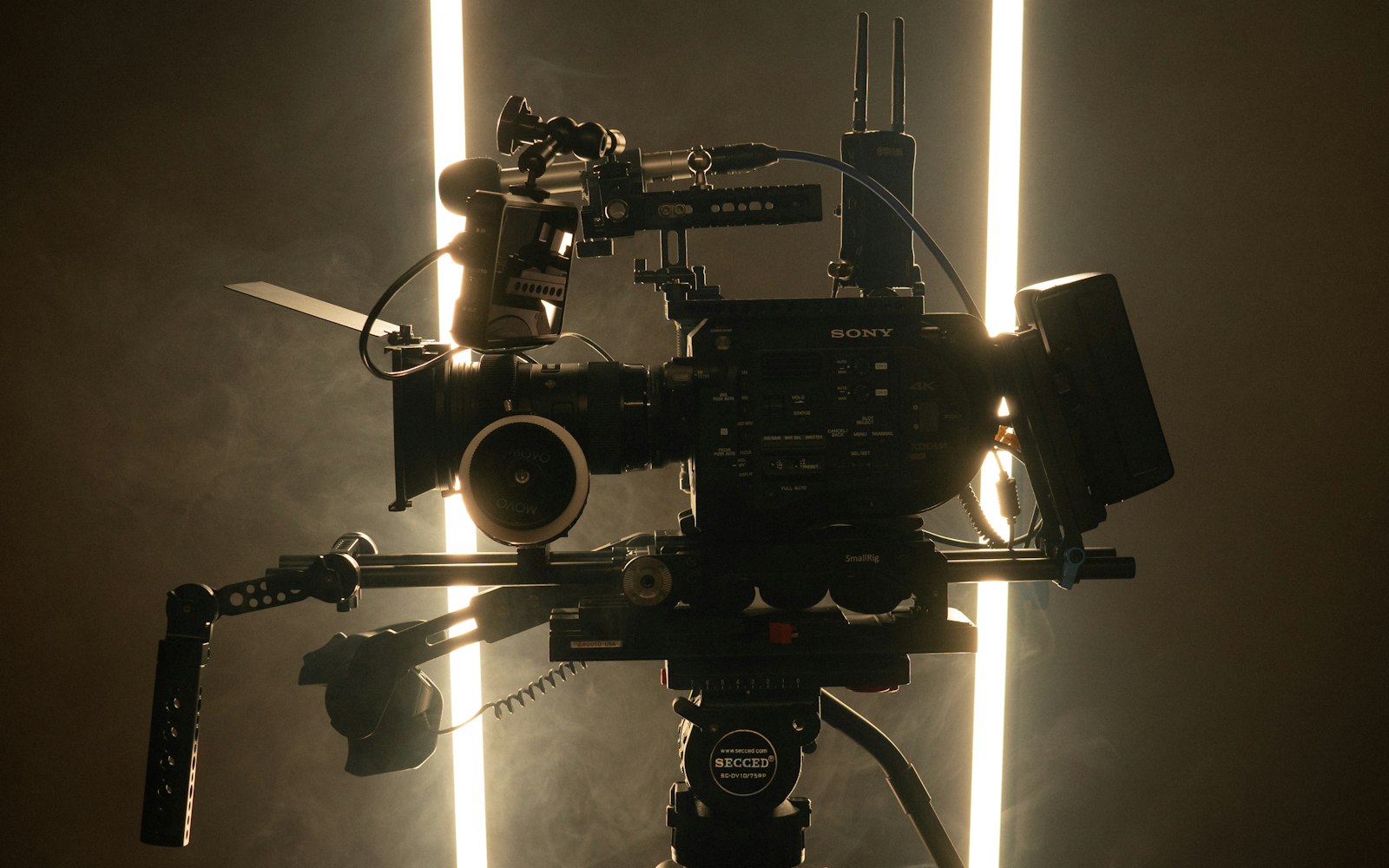 A rigged cinema camera silhouetted against two light tubes in haze