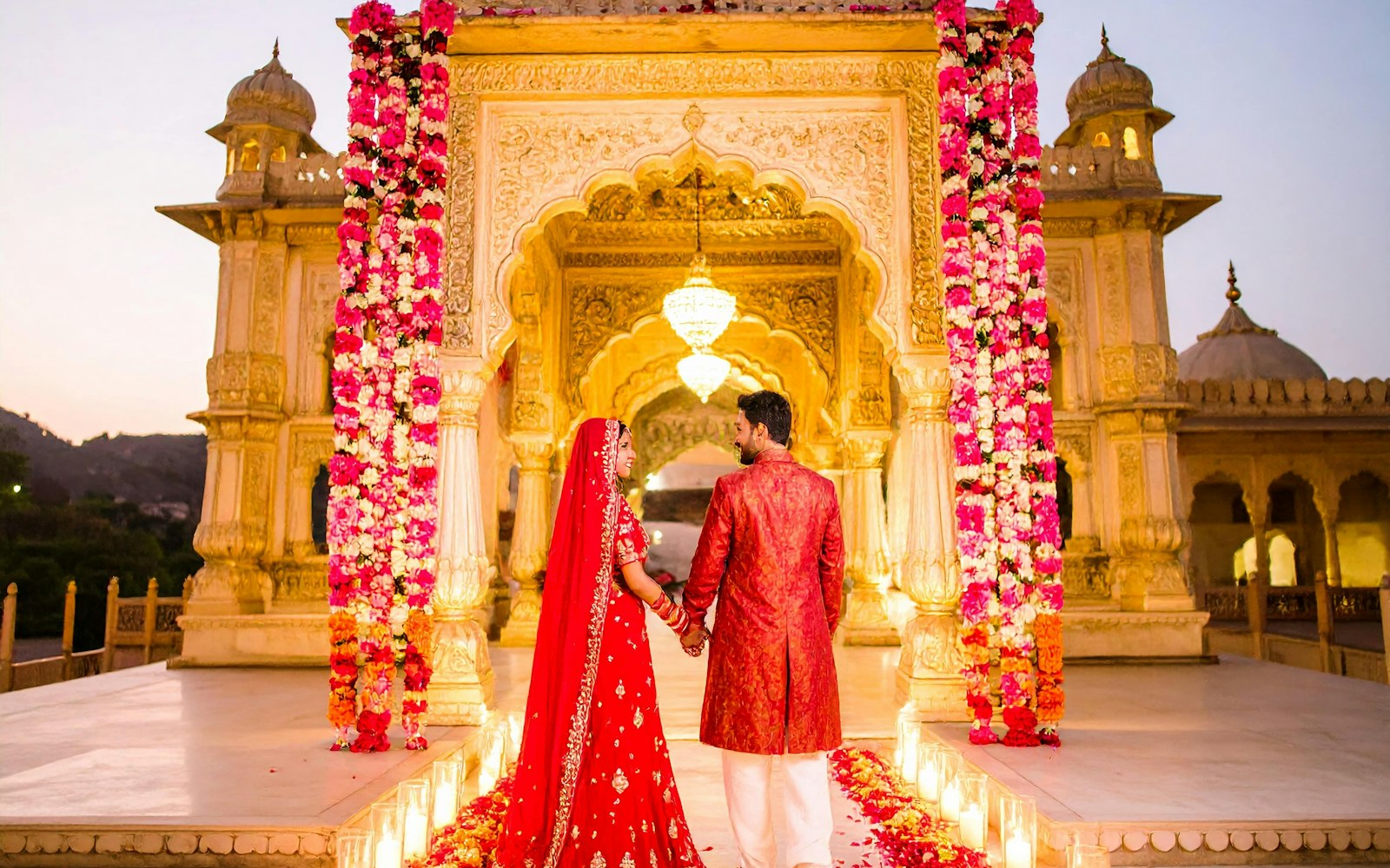 A bride and groom in red walking toward a palace pavilion framed by flower columns